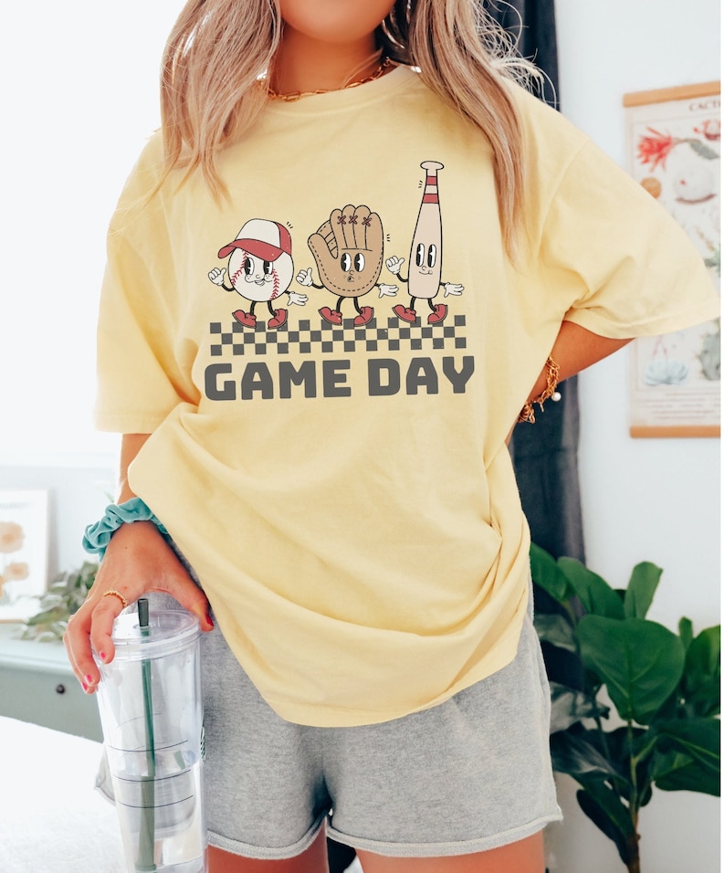 May include: A yellow t-shirt featuring a graphic of a baseball, glove, and bat with faces. The graphic is in a retro style and says "GAME DAY" underneath.