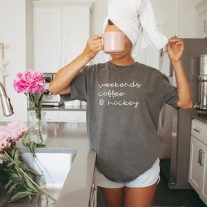 May include: A person wearing a gray t-shirt with the text "weekends coffee & hockey" and gray shorts, holding a pink mug. A white towel is wrapped around their head. Pink peonies are in a vase on the counter.