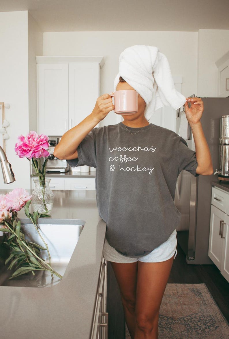 May include: A person wearing a gray t-shirt with the text "weekends coffee & hockey" and gray shorts, holding a pink mug. A white towel is wrapped around their head. Pink peonies are in a vase on the counter.
