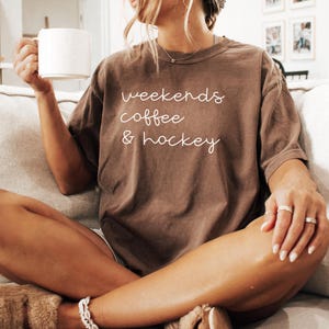 May include: A brown t-shirt with the words "weekends coffee & hockey" in white script. The person is holding a white mug. The person is wearing rings and a bracelet.