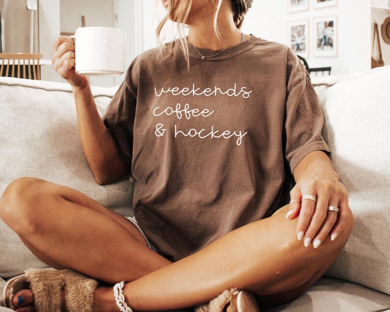 May include: A brown t-shirt with the words "weekends coffee & hockey" in white script. The person is holding a white mug. The person is wearing rings and a bracelet.