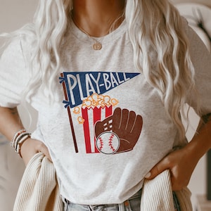 May include: A gray t-shirt with a graphic of a baseball glove, popcorn, and a pennant that says "PLAY-BALL".