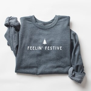 Op de afbeelding: Een donkergrijs sweatshirt met de tekst "FEELIN' FESTIVE" en een kleine witte kerstboomgrafiek.