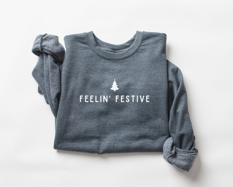 Op de afbeelding: Een donkergrijs sweatshirt met de tekst "FEELIN' FESTIVE" en een kleine witte kerstboomgrafiek.