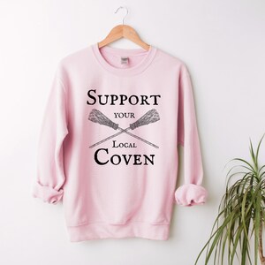 May include: Pink sweatshirt with the text "Support Your Local Coven" and a graphic of two crossed brooms.