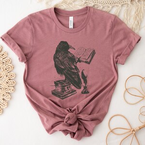 Puede incluir: Una camiseta color malva con una ilustraci&oacute;n en blanco y negro de un cuervo leyendo un libro. El cuervo est&aacute; posado sobre una pila de libros con una pluma y un tintero a su lado.