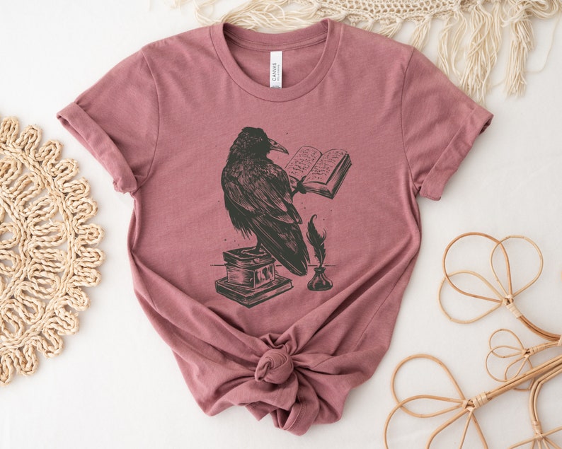 Puede incluir: Una camiseta color malva con una ilustraci&oacute;n en blanco y negro de un cuervo leyendo un libro. El cuervo est&aacute; posado sobre una pila de libros con una pluma y un tintero a su lado.