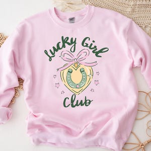 May include: Pink sweatshirt with the text "Lucky Girl Club" in green and black lettering. A green horseshoe with a pink bow is in the center of the design.