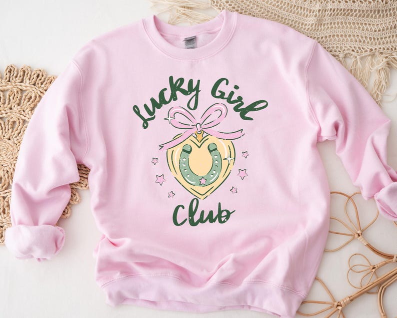 May include: Pink sweatshirt with the text "Lucky Girl Club" in green and black lettering. A green horseshoe with a pink bow is in the center of the design.