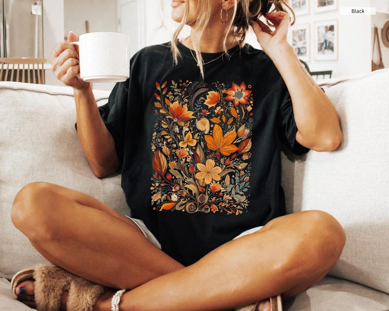 May include: Black t-shirt featuring a vibrant floral design in shades of orange, yellow, and brown. The botanical illustration includes various flowers and leaves. The model is holding a white mug. The shirt is a comfortable, casual style.