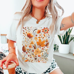May include: White t-shirt with a floral print featuring yellow, orange, and brown flowers and leaves.