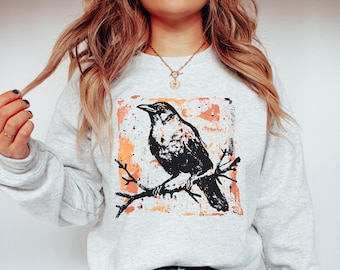 May include: A light gray sweatshirt featuring a black and white graphic of a crow perched on a branch. The graphic is set against a textured orange and white background. The sweatshirt has long sleeves and a crew neck.