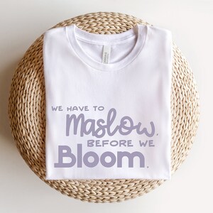 May include: White t-shirt with the text "We have to Maslow before we Bloom." in purple lettering.