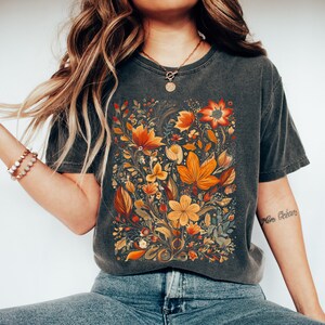 May include: Dark gray t-shirt featuring a vibrant floral design in shades of orange, yellow, and red. The botanical illustration includes various flowers and leaves, creating a detailed and colorful pattern. The shirt has a classic crew neck and short sleeves.