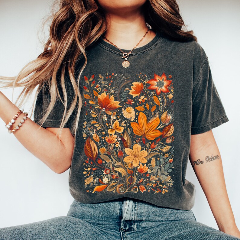 May include: Dark gray t-shirt featuring a vibrant floral design in shades of orange, yellow, and red. The botanical illustration includes various flowers and leaves, creating a detailed and colorful pattern. The shirt has a classic crew neck and short sleeves.