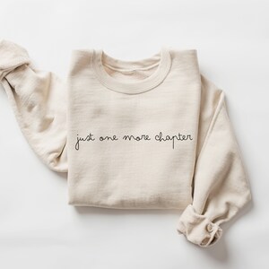 May include: A beige sweatshirt with the text "just one more chapter" in black script.