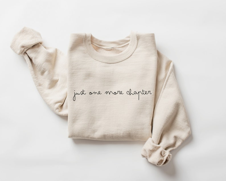 May include: A beige sweatshirt with the text "just one more chapter" in black script.