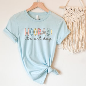 May include: A light blue t-shirt with the text "HOORAY! it's art day" in colourful letters. The shirt is tied in a knot at the bottom.
