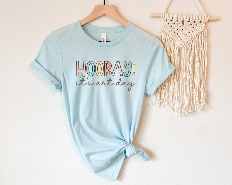 May include: A light blue t-shirt with the text "HOORAY! it's art day" in colourful letters. The shirt is tied in a knot at the bottom.