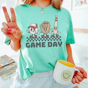 May include: A teal blue t-shirt featuring a graphic of a baseball, glove, and bat with faces. The graphic is in a retro style and the text "GAME DAY" is printed below it.