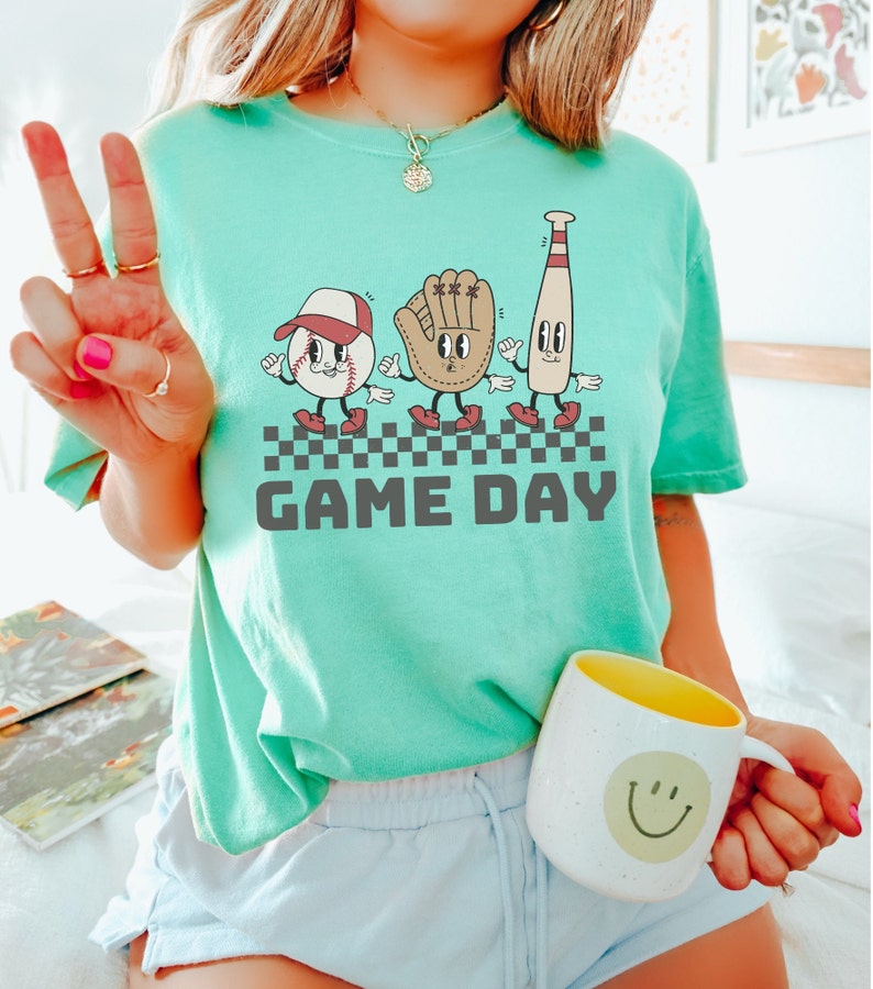 May include: A teal blue t-shirt featuring a graphic of a baseball, glove, and bat with faces. The graphic is in a retro style and the text "GAME DAY" is printed below it.
