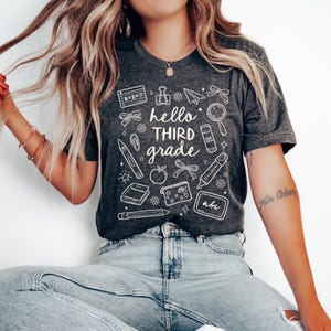 May include: Dark gray t-shirt with the words "hello THIRD grade" in white, surrounded by school-themed illustrations. The design includes a calculator, paperclip, pencil, apple, and other school supplies.