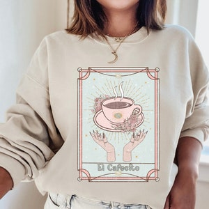 May include: A beige sweatshirt with a graphic print of a coffee cup with steam rising from it, surrounded by flowers and stars. The graphic is in a pink and blue color scheme and has the text "El Cafeito" at the bottom.