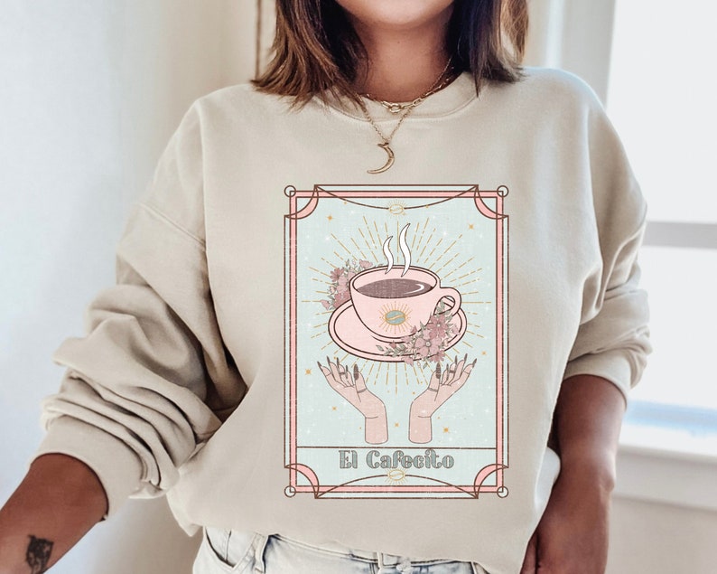 May include: A beige sweatshirt with a graphic print of a coffee cup with steam rising from it, surrounded by flowers and stars. The graphic is in a pink and blue color scheme and has the text "El Cafeito" at the bottom.