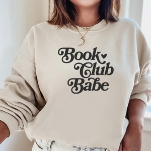 K&ouml;nnte beinhalten: Ein beiger Pullover mit dem Text "Book Club Babe" in einer Retro-Schrift.
