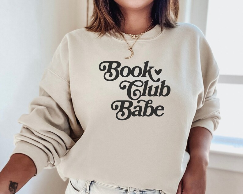 K&ouml;nnte beinhalten: Ein beiger Pullover mit dem Text "Book Club Babe" in einer Retro-Schrift.