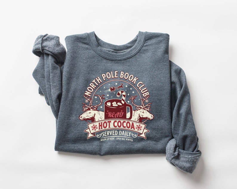 Grey crewneck with 'North Pole Book club hot cocoa served daily' in a red and white Christmas graphic with reindeer, a cup of hot cocoa that says 'read' with marshmallows and a candy cane in it.
