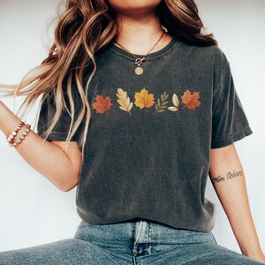 May include: A dark gray t-shirt with a row of colorful autumn leaves printed on the front. The leaves are in shades of red, orange, yellow, and green.