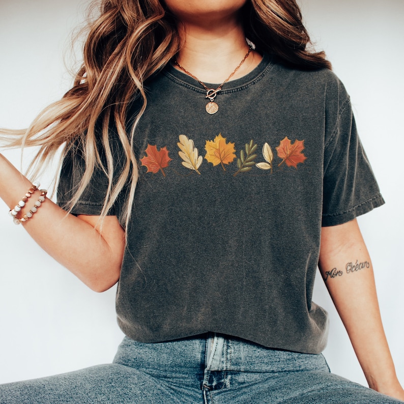 May include: A dark gray t-shirt with a row of colorful autumn leaves printed on the front. The leaves are in shades of red, orange, yellow, and green.