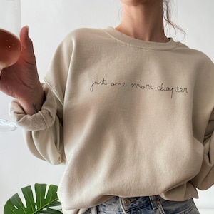 May include: A beige sweatshirt with the text "just one more chapter" in black script.
