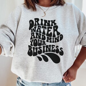 May include: Gray sweatshirt with a black graphic print that says "Drink Water And Mind Your Business" in a retro font.