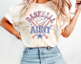 May include: A cream-colored short-sleeve t-shirt featuring a graphic print with the text BASEBALL AUNT. The design includes two crossed baseball bats, a baseball, and a star, all rendered in a distressed red, white, and blue color palette.