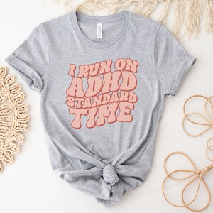 May include: Grey t-shirt with the text "I run on ADHD standard time" in pink retro-style lettering.