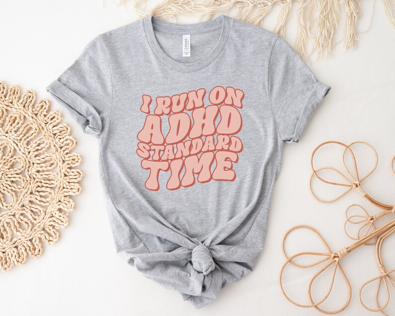 May include: Grey t-shirt with the text "I run on ADHD standard time" in pink retro-style lettering.