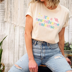 Pu&ograve; includere: Una donna indossa una maglietta beige chiaro con il testo "It's a neurodiverse universe" in colori arcobaleno e una grafica di un pianeta rosa. Indossa jeans blu con dettagli usurati.