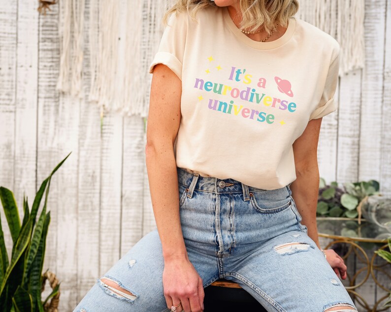 Pu&ograve; includere: Una donna indossa una maglietta beige chiaro con il testo "It's a neurodiverse universe" in colori arcobaleno e una grafica di un pianeta rosa. Indossa jeans blu con dettagli usurati.