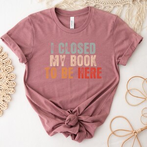 Puede incluir: Una camiseta color malva con el texto "I CLOSED MY BOOK TO BE HERE" en un esquema de colores arco&iacute;ris retro.