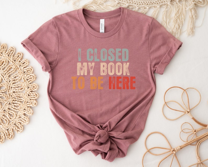 Puede incluir: Una camiseta color malva con el texto "I CLOSED MY BOOK TO BE HERE" en un esquema de colores arco&iacute;ris retro.