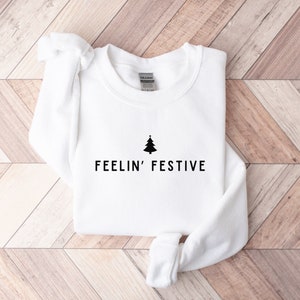 Op de afbeelding: Witte trui met een zwarte kerstboom en de tekst "FEELIN' FESTIVE" bedrukt op de voorkant.