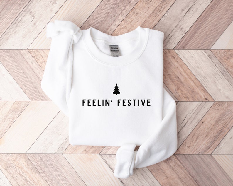 Op de afbeelding: Witte trui met een zwarte kerstboom en de tekst "FEELIN' FESTIVE" bedrukt op de voorkant.