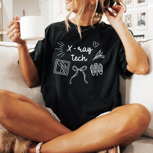 K&ouml;nnte beinhalten: Ein schwarzes T-Shirt mit wei&szlig;em Text, der "X-ray tech" lautet und Illustrationen einer R&ouml;ntgenaufnahme eines Knies, einer Schleife und einer R&ouml;ntgenaufnahme eines Brustkorbs enth&auml;lt.