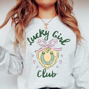 May include: A gray sweatshirt with the text "Lucky Girl Club" printed on the front. The text is in green and black. A pink and green horseshoe charm is centered below the text.