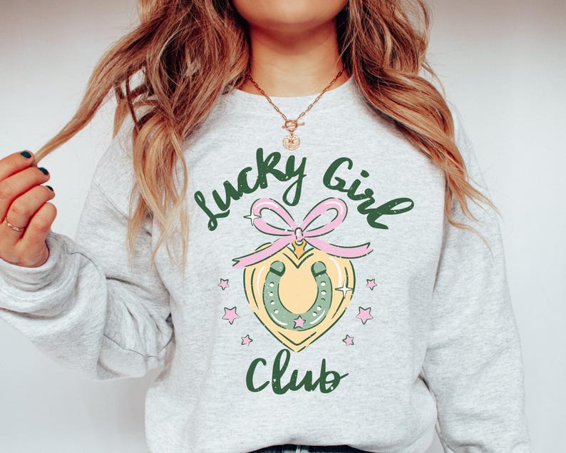 May include: A gray sweatshirt with the text "Lucky Girl Club" printed on the front. The text is in green and black. A pink and green horseshoe charm is centered below the text.