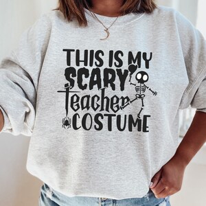 May include: A light gray sweatshirt with the text "THIS IS MY SCARY TEACHER COSTUME" in black. A skeleton graphic is included. A spider graphic is also present. The sweatshirt is a Halloween-themed item.