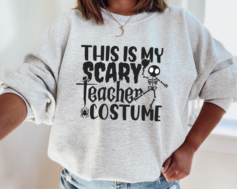 May include: A light gray sweatshirt with the text "THIS IS MY SCARY TEACHER COSTUME" in black. A skeleton graphic is included. A spider graphic is also present. The sweatshirt is a Halloween-themed item.