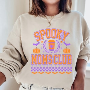 May include: A beige sweatshirt with a purple and orange graphic design that says "Spooky Moms Club". The graphic includes a coffee cup with a spider, pumpkins, bats, and a spiderweb.